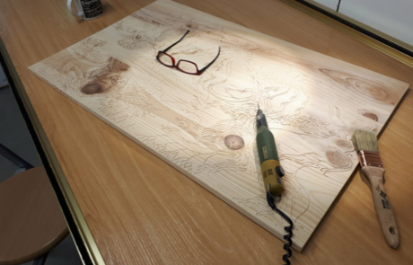 Detalle de una tabla de madera preparada para ser tallada. Sobre la tabla hay un cincel electrónico, un pincel y unas gafas. Al fondo de la mesa se ve una taza de té.
