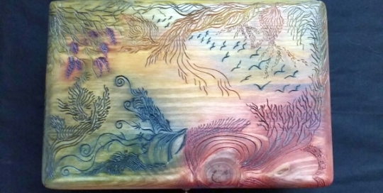 Caja de madera de pino con la tapa tallada con simbología de Samu-Do. Evoca un paisaje paradisíaco con diversa vegetación y una bandada de pájaros volando en el cielo