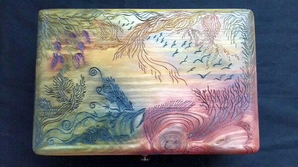 Caja de madera de pino con la tapa tallada con simbología de Samu-Do. Muestra una una bandada de pájaros volando en el cielo vistos desde un lugar con mucha vegetación